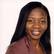 Kiki, a black woman with hair past shoulder length wearing a brown blazer and a light pink shirt underneath.